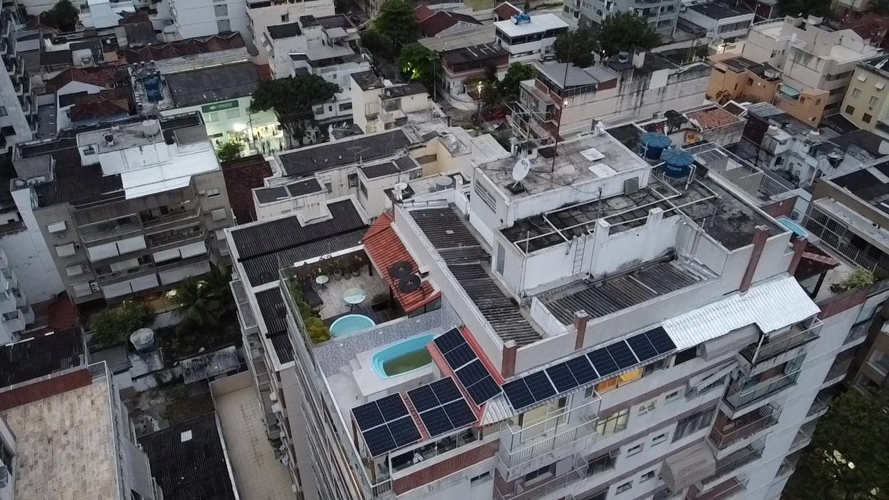 Vista aérea de prédio residencial denso com painéis solares e piscina na cobertura, ao entardecer